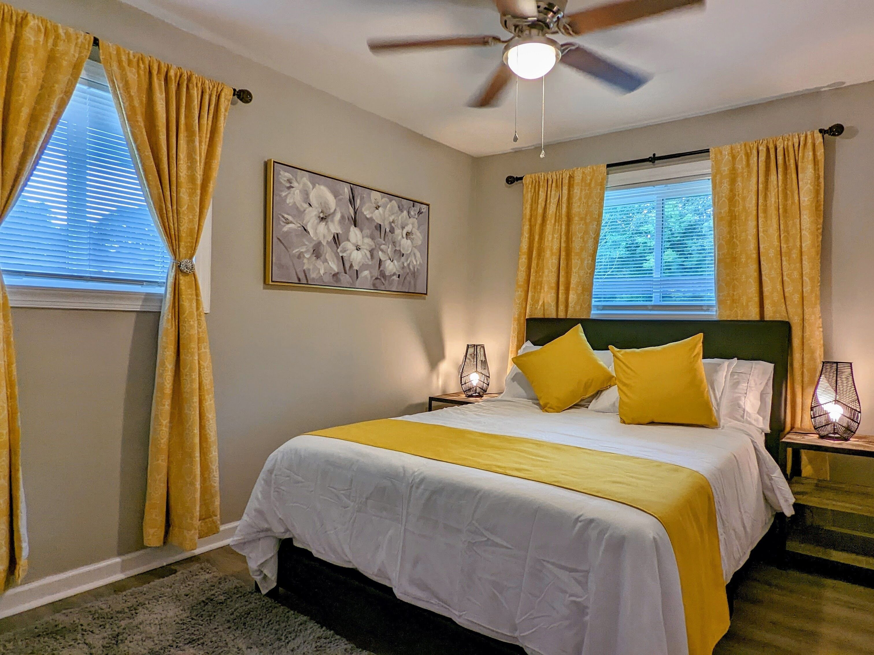 A bedroom with a bed, curtains, a ceiling fan, a nightstand, and a wall-mounted lamp.