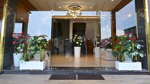 Ingresso moderno di un hotel con porte in vetro, un lampadario centrale e piante in vaso.