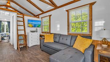 Ein gemütliches Zimmer mit grauem Sofa, Holzbalken, einer Leiter und einem Fernseher an der Wand.