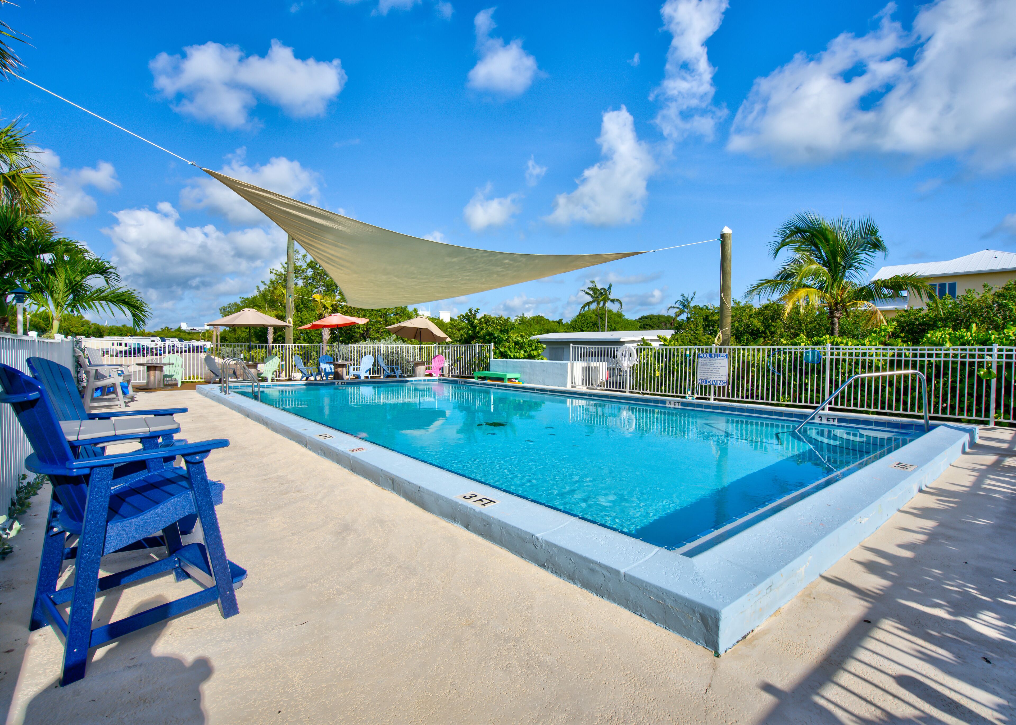A rectangular swimming pool with a concrete edge, surrounded by a white metal fence. There are several lounge chairs, some with umbrellas, and a few colorful chairs. A large canopy provides shade over part of the pool area.