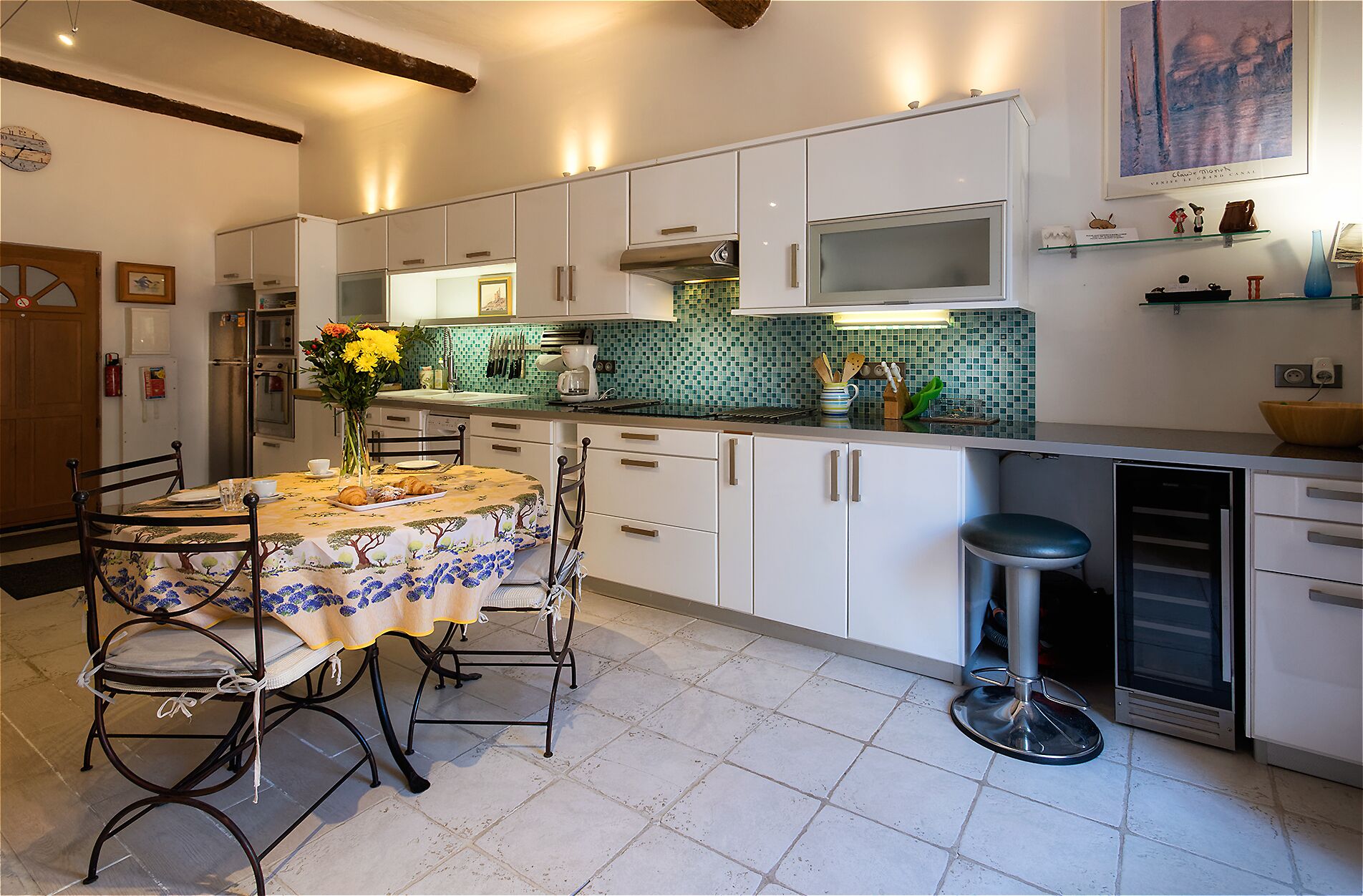 Une cuisine avec des meubles blancs, un pare-feu en carrelage, une table à manger avec des chaises et un tabouret de bar.