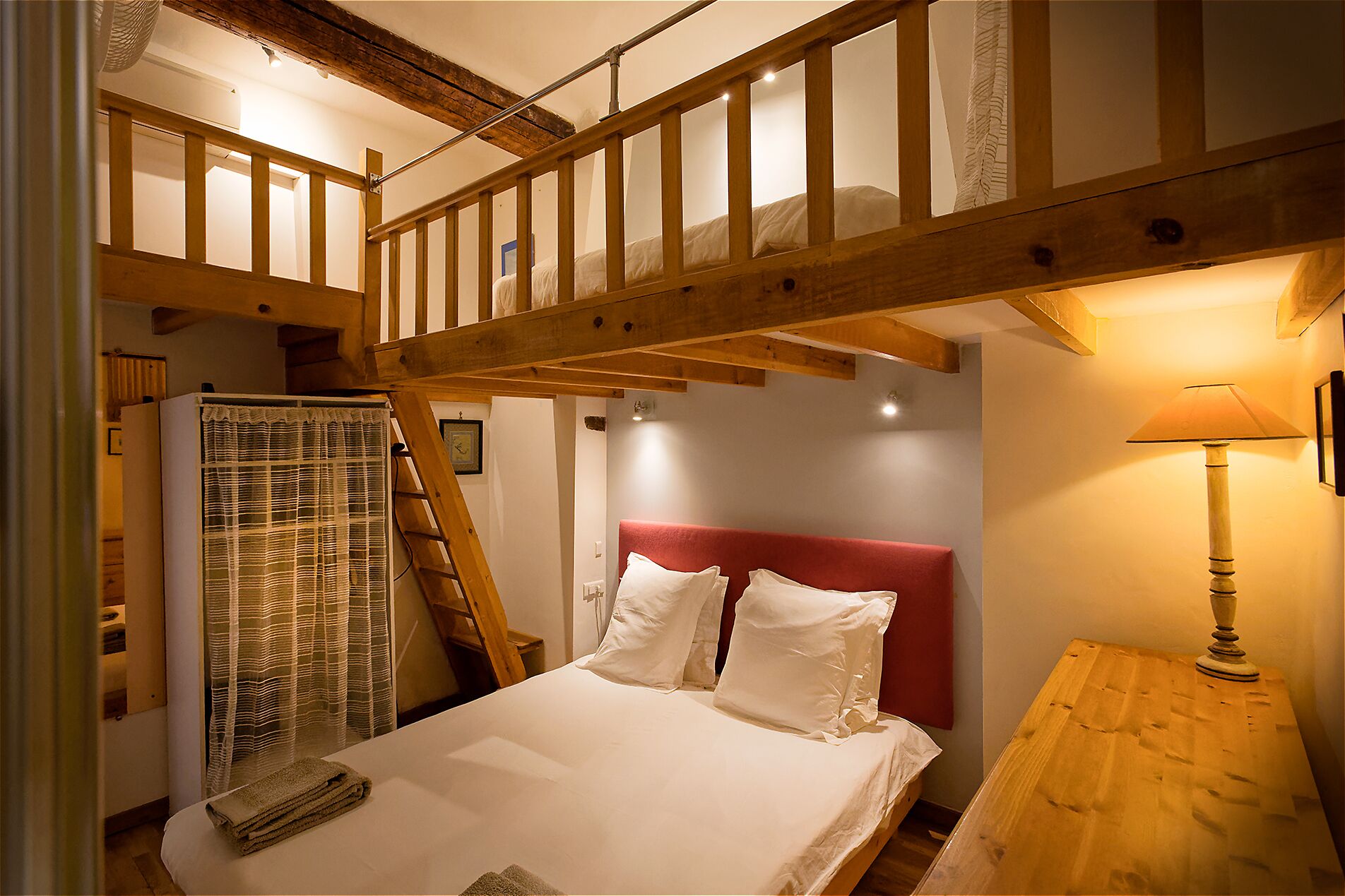 Une chambre avec un escalier en bois, un lit avec une literie blanche, une tête de lit rouge, une table de chevet en bois et une lampe.
