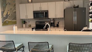 Una cocina moderna con armarios blancos, un refrigerador de acero inoxidable, un microondas y una cafetera. Hay tres sillas negras de malla metálica en la isla.