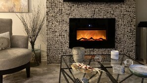 Una sala de estar con chimenea de piedra, un televisor de pantalla plana montado arriba, un sillón cómodo, una mesa de centro de vidrio con objetos decorativos y un jarrón con ramas.