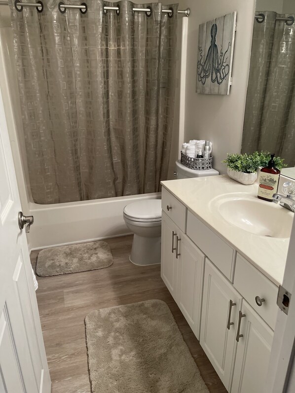 Baño con bañera, inodoro y lavamanos. Hay una cortina de ducha, un perchero y una planta en maceta.