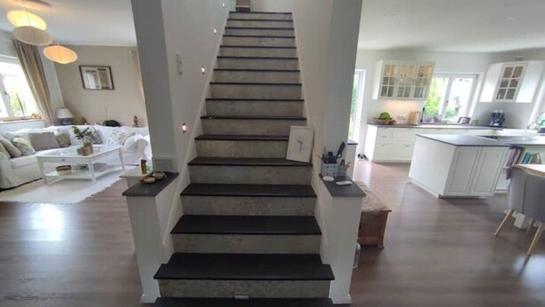Eine Treppe führt zu einer zweiten Etage, umgeben von einem Wohnbereich mit einer Couch und einem Couchtisch sowie einer Küchenzeile mit Schränken und einer Arbeitsplatte.