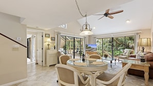 A spacious living area with a round glass table, wicker chairs, a ceiling fan, and a large window offering a view of the outdoors. - SeaPines, On-Site Pool and Tennis, Screened Porch, Pet Friendly (Hilton Head Island)