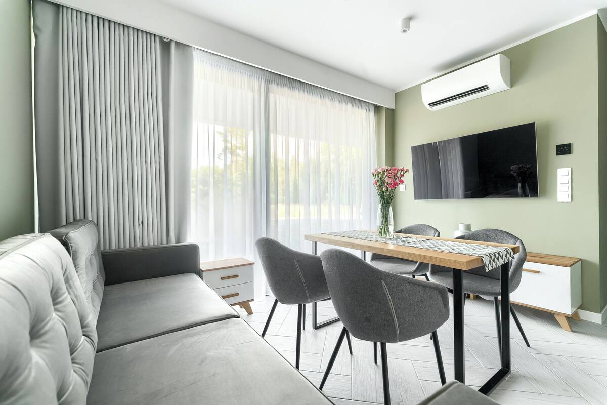 A modern living room with a sofa, dining table, chairs, a flat-screen TV, and a large window with curtains.