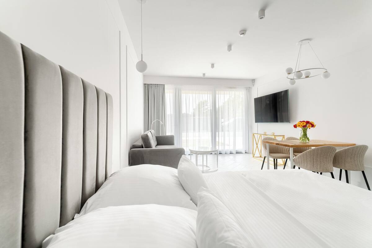 A modern hotel room with a large bed, a dining area with a table and chairs, and a TV mounted on the wall.
