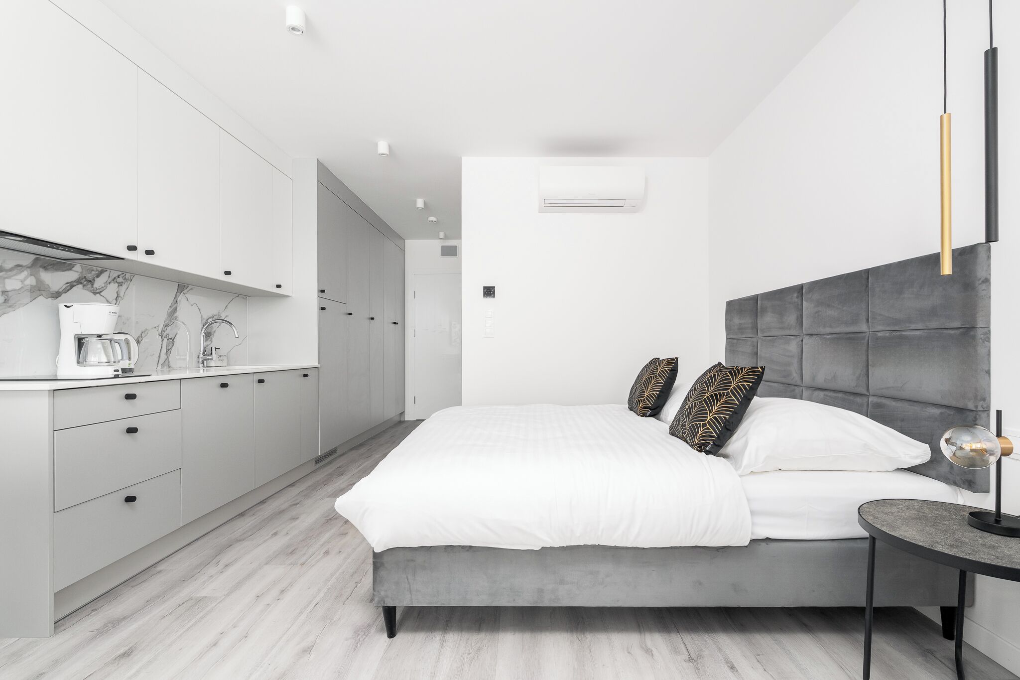 A modern bedroom with a grey upholstered headboard, a white bedspread, and a bedside table with a lamp.
