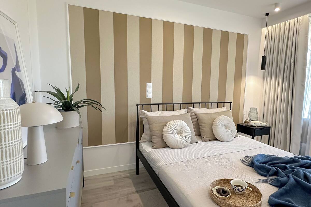 A neatly arranged bedroom with a striped headboard, a bedside table with a lamp, a potted plant, and a cozy blanket on the bed.