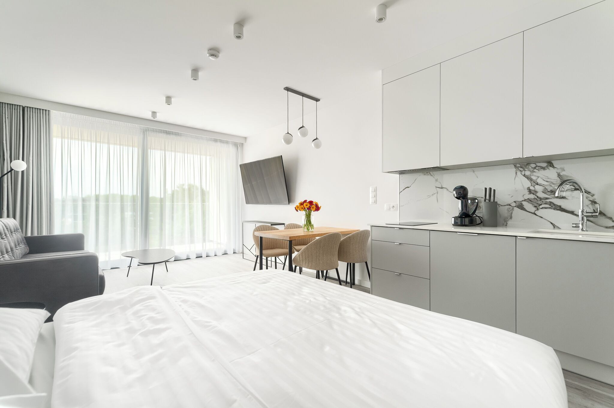 A modern hotel room with a large bed, a dining area with a table and chairs, a kitchen area with a marble countertop, and a TV mounted on the wall.