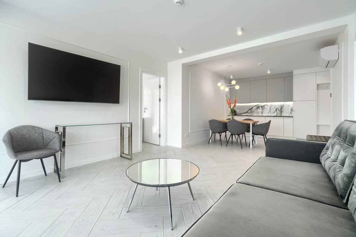 A modern living room with a grey sofa, a round glass coffee table, and a grey armchair. There is a flat-screen TV mounted on the wall, a dining area with a table and chairs, and a kitchen visible through an open doorway.