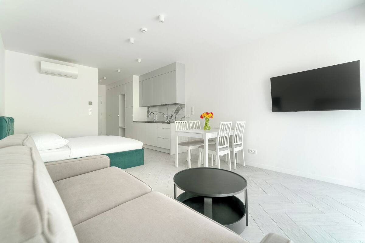 A modern living room with a sofa, a dining table with chairs, a flat-screen TV mounted on the wall, and a kitchen area in the background.