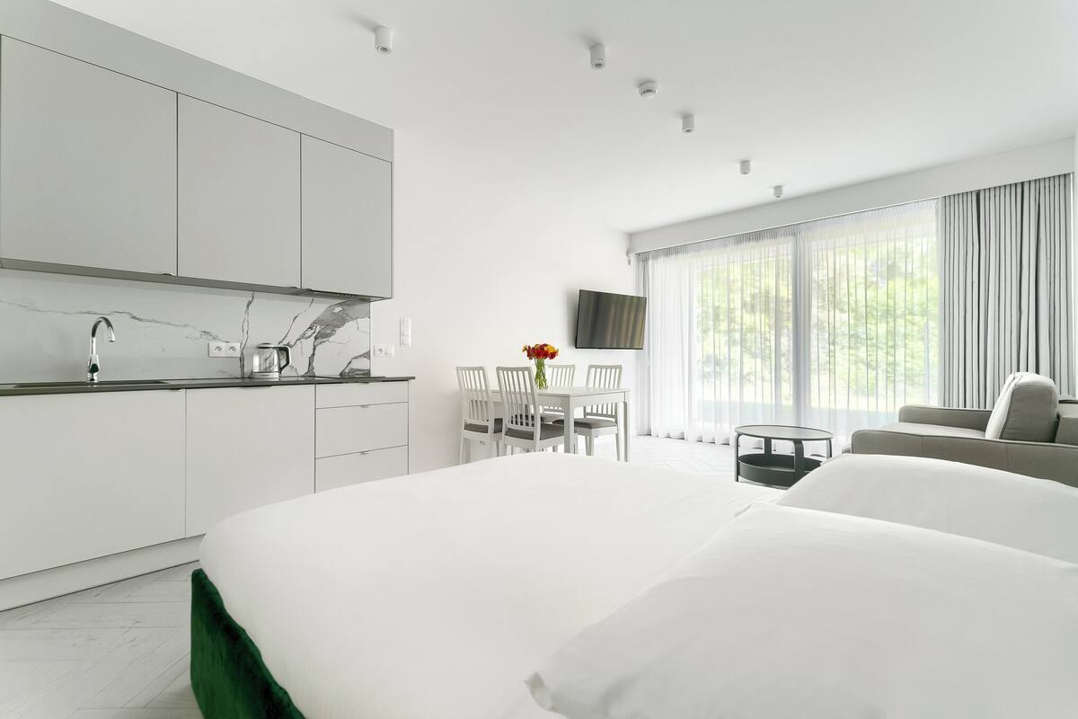 A modern hotel room with a large bed, a dining area with a table and chairs, a kitchen with white cabinets, and a large window with curtains.