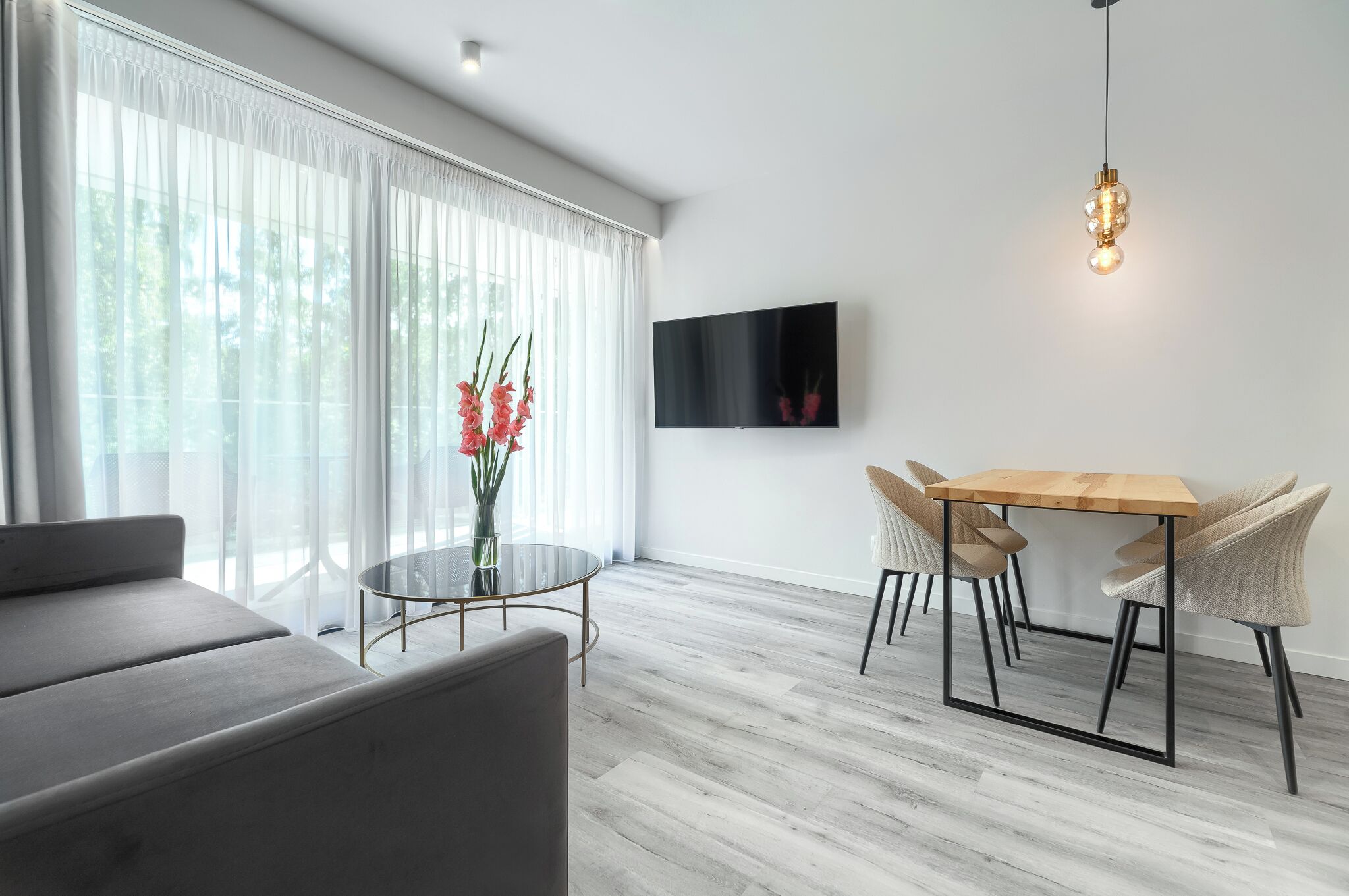 A modern living room with a sofa, a round glass table, and a dining table with chairs. There is a TV mounted on the wall and a hanging light fixture.