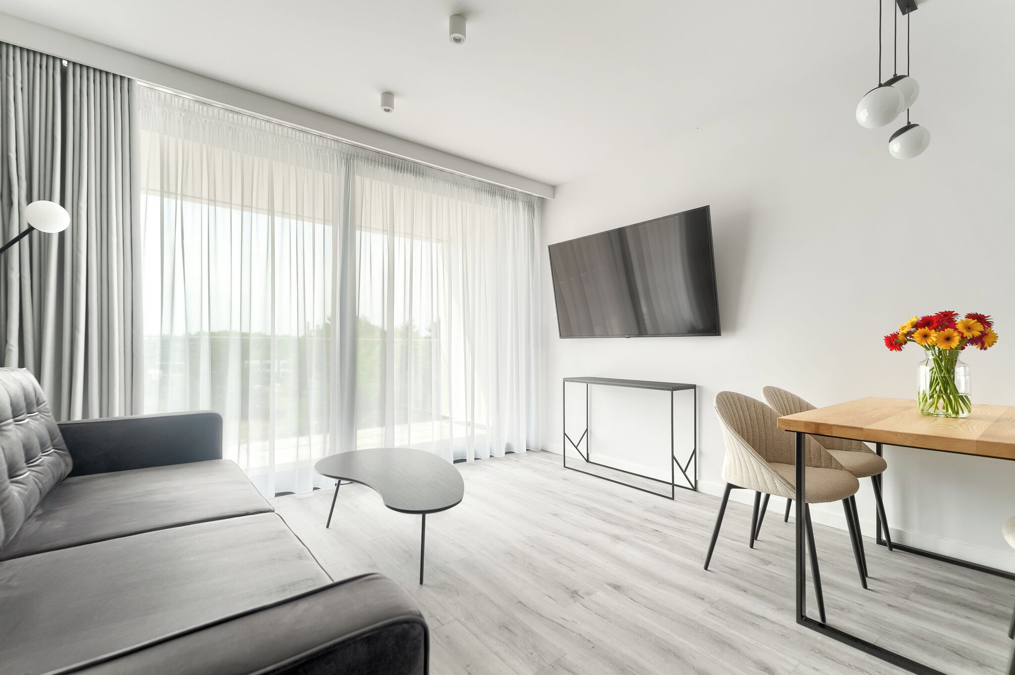 Una sala de estar moderna con un sofá, una mesa de comedor con sillas, un televisor de pantalla plana y un amplio ventanal con cortinas translúcidas.
