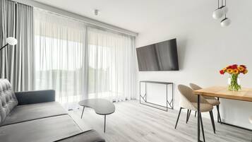 Una sala de estar moderna con un sofá, una mesa de comedor con sillas, un televisor de pantalla plana y un amplio ventanal con cortinas translúcidas.
