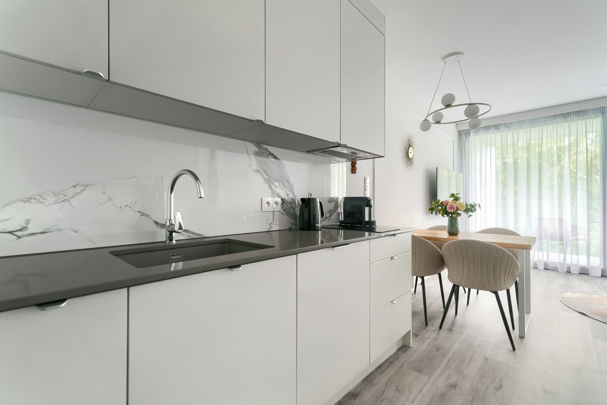 Una cocina moderna con armarios blancos, salpicadero de mármol y encimera negra. Hay una cafetera sobre la encimera y un comedor con mesa y sillas.