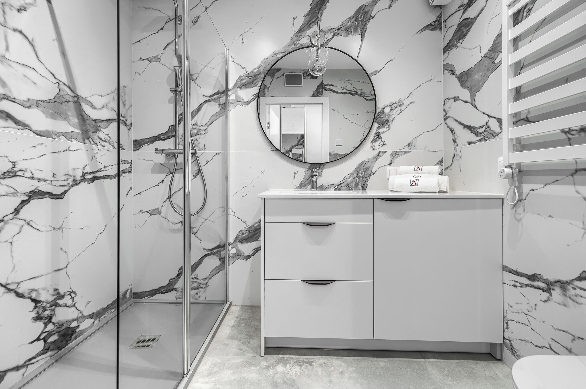 Une salle de bain moderne avec des murs en marbre, une douche en verre, un meuble-lavabo blanc avec des tiroirs et un miroir rond.