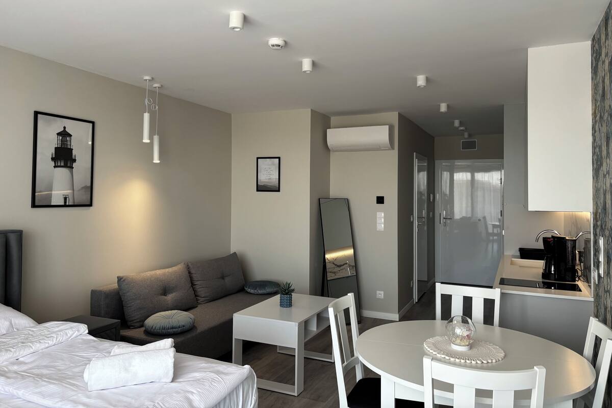 A modern hotel room with a bed, a sofa, a dining table with chairs, a small kitchen area, and a bathroom visible through an open door.