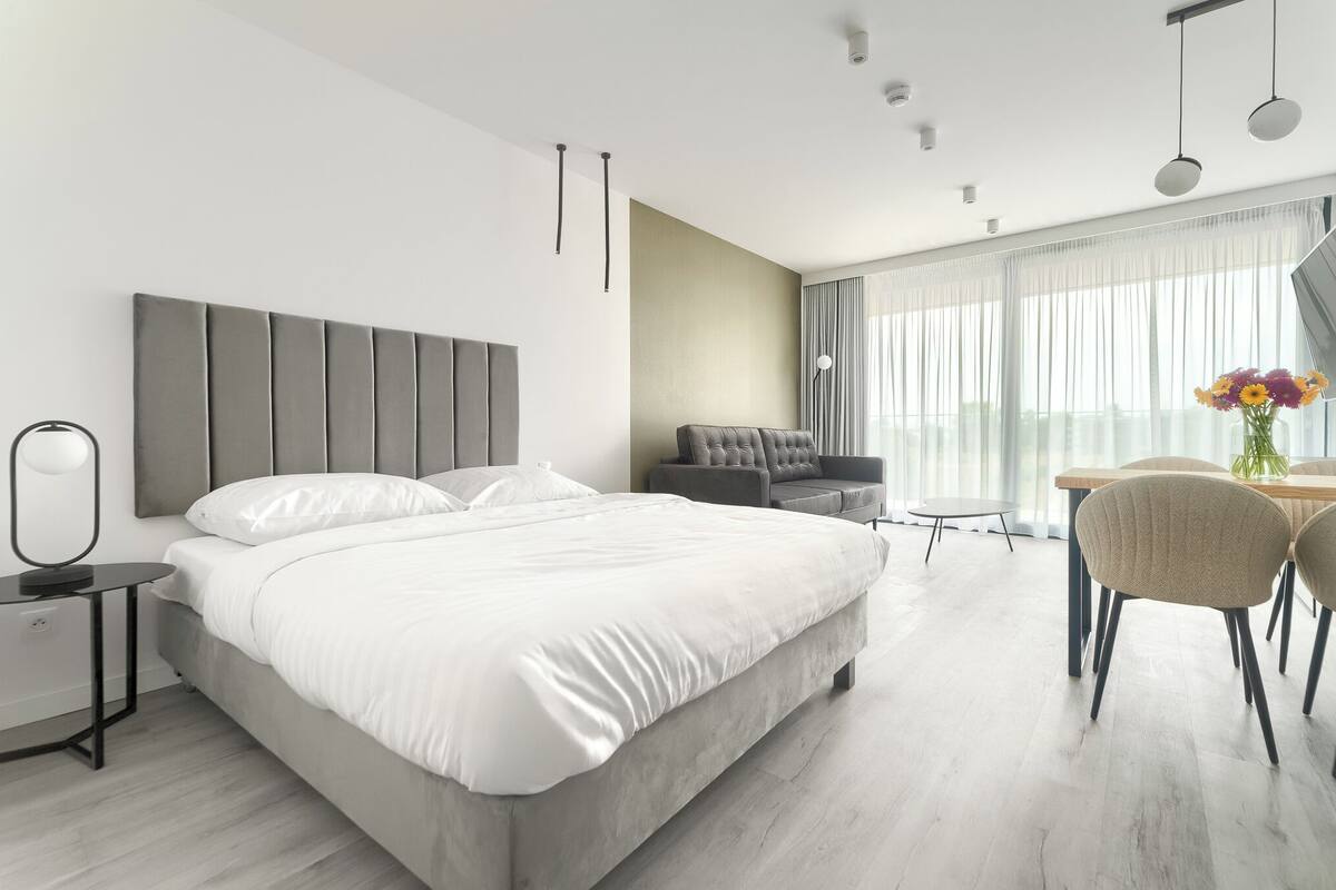 A modern hotel room with a large bed, a desk with chairs, and a view of the outdoors.