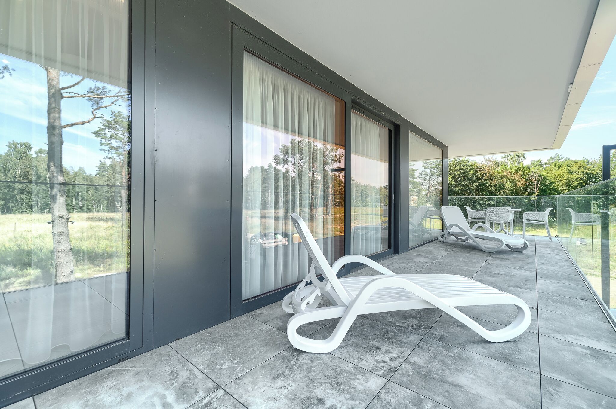 A modern outdoor balcony with a glass door, white lounge chairs, and a view of a grassy area.
