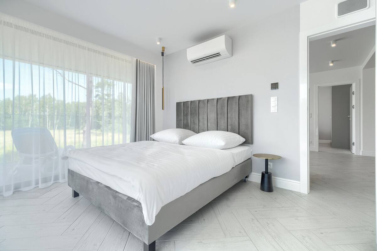A modern bedroom with a large bed, a bedside table, and a view of the outdoors through sheer curtains.