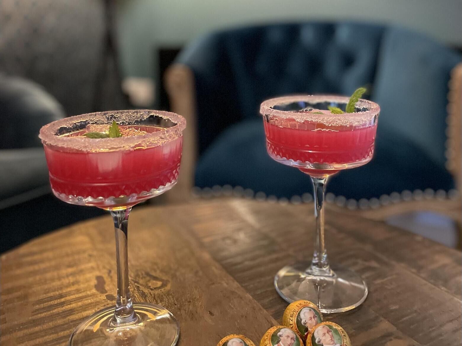 Two cocktails on a wooden table with a blue chair in the background.