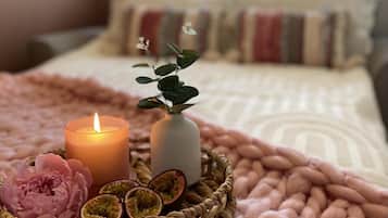 A cozy bedroom with a bed, a lit candle, a vase with a plant, and a basket with flowers and passion fruit halves.