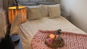 Una camera da letto ordinata con un letto, cuscini, una lampada da comodino, un vaso con piume e un vassoio con candele e una pianta.