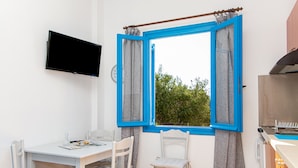 Une pièce comprenant une table blanche, deux chaises blanches, un téléviseur à écran plat, une fenêtre avec des cadres bleus et un coin cuisine équipé d’un gril.
