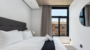 A modern hotel room with a large bed, a headboard, a window with curtains, and a view of a brick building. - The Signature G07 (Cape Town)