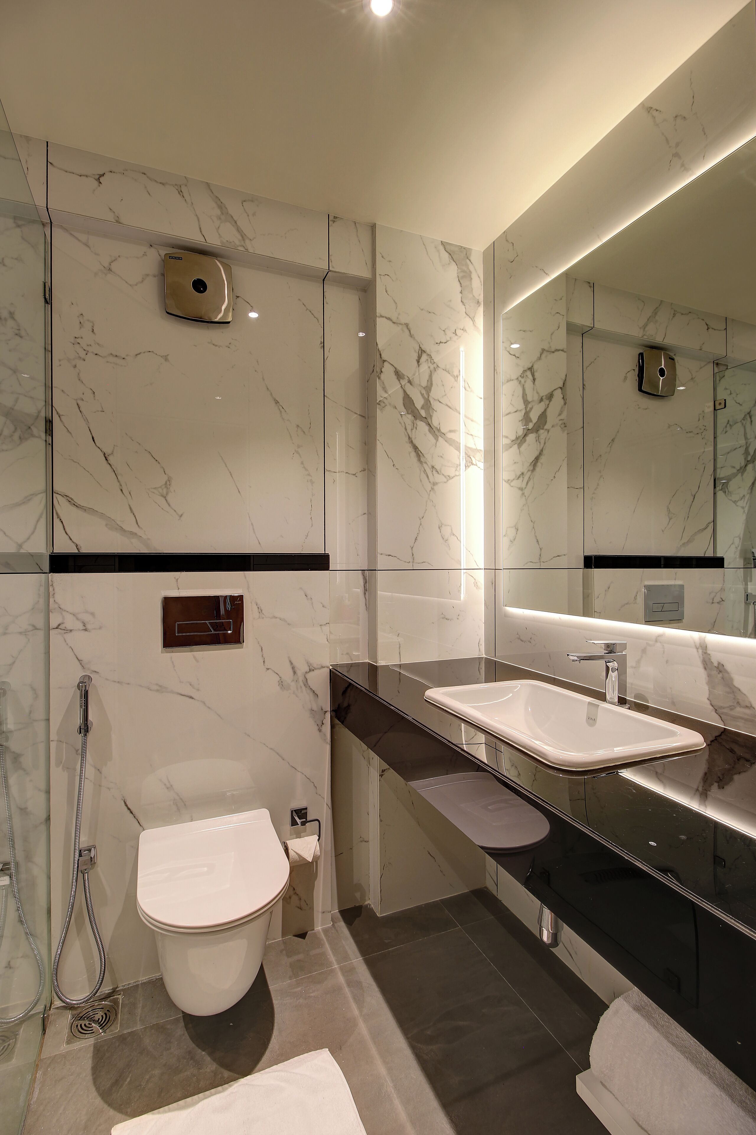 Ein modernes Badezimmer mit Marmorwänden, einem großen Spiegel, einer weißen Toilette und einer schwarzen Ablagefläche mit Waschbecken.