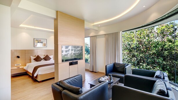 Una habitación de hotel moderna con cama, televisión, zona de estar con mesa y un amplio ventanal con vista a zonas verdes.