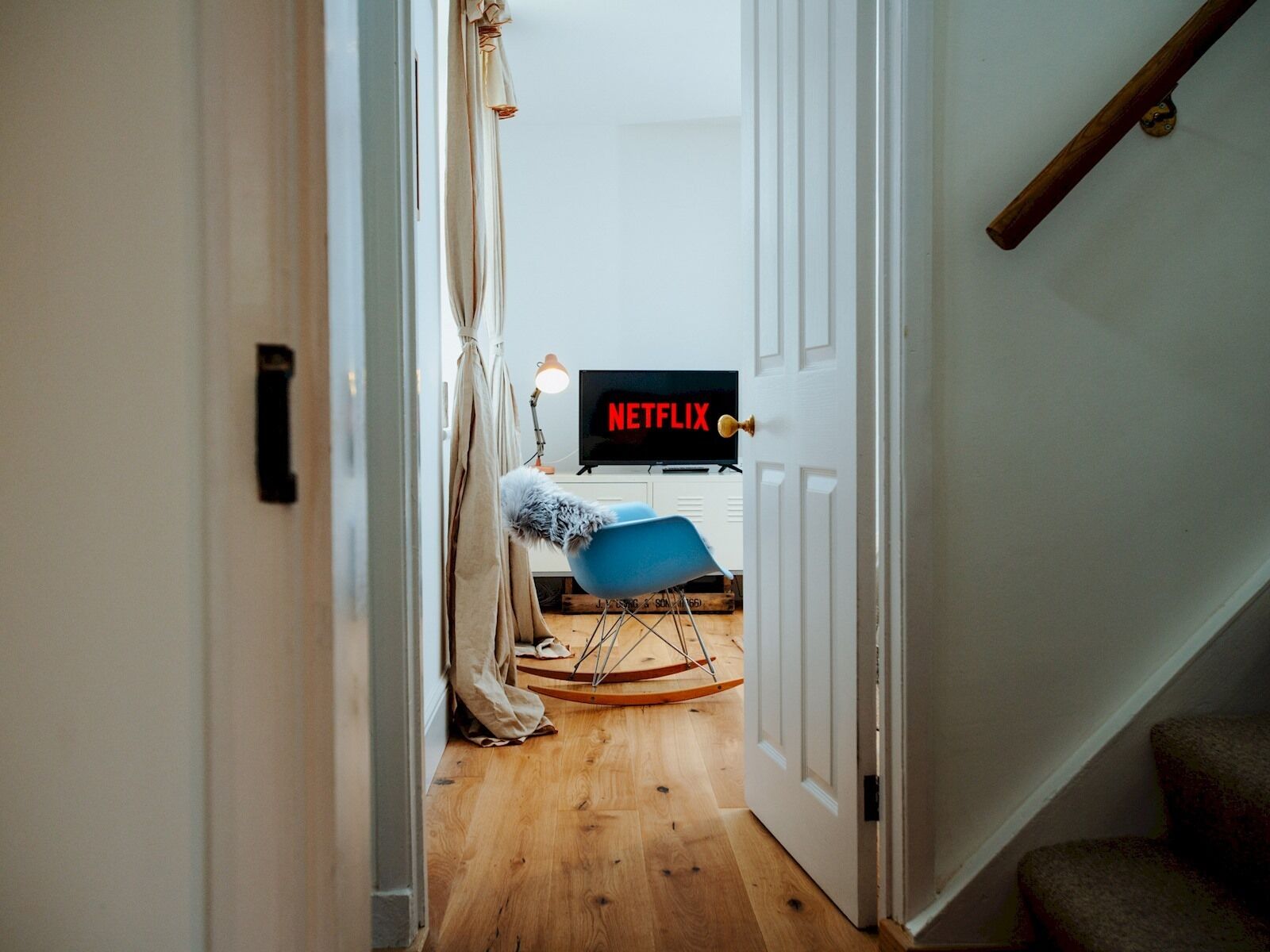 A room with a wooden floor, a blue chair, a television displaying 'NETFLIX', and a door leading to another room.