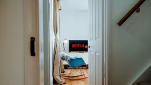 Huoneessa on puulattia, sininen tuoli, televisio, jossa näkyy teksti ”NETFLIX”, ja ovi, joka johtaa toiseen huoneeseen.
