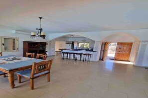 Uma ampla área de refeições com mesa e cadeiras de madeira, uma cozinha ao fundo e uma porta em arco que dá para o exterior.