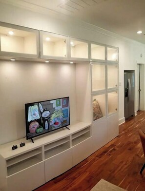 A modern living room with a flat-screen TV on a white entertainment unit, wooden flooring, and built-in shelving with recessed lighting.