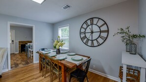 Un coin repas avec une table en bois dressée pour quatre personnes, une grande horloge murale et une fenêtre équipée de stores.