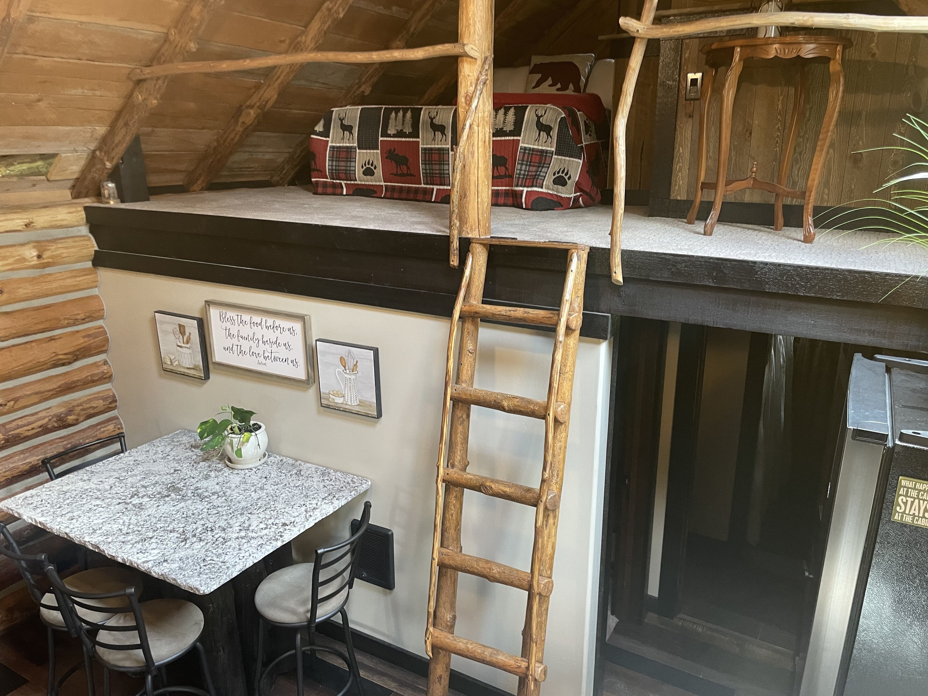 A rustic cabin interior with a wooden ladder leading to a lofted sleeping area. A small table with chairs is positioned under the loft, and a plant is on the table.