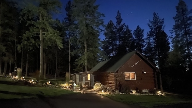 A log cabin with a porch, surrounded by trees, and illuminated by ground lights. - Little Cabin in the Woods + Disk - Frisbee Golf (Post Falls)
