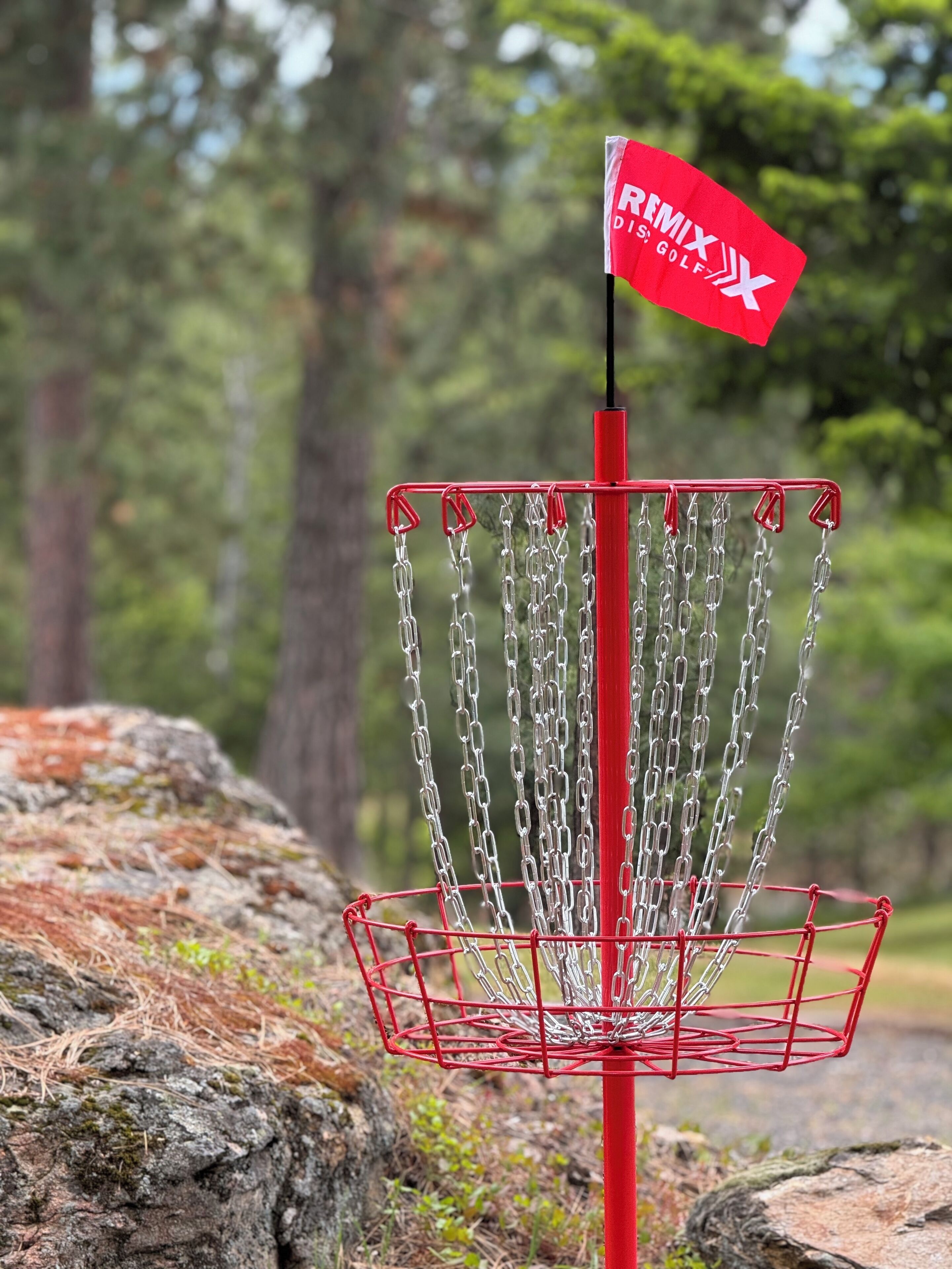A disc golf basket with a red flag on top, surrounded by chains and set against a forest backdrop.