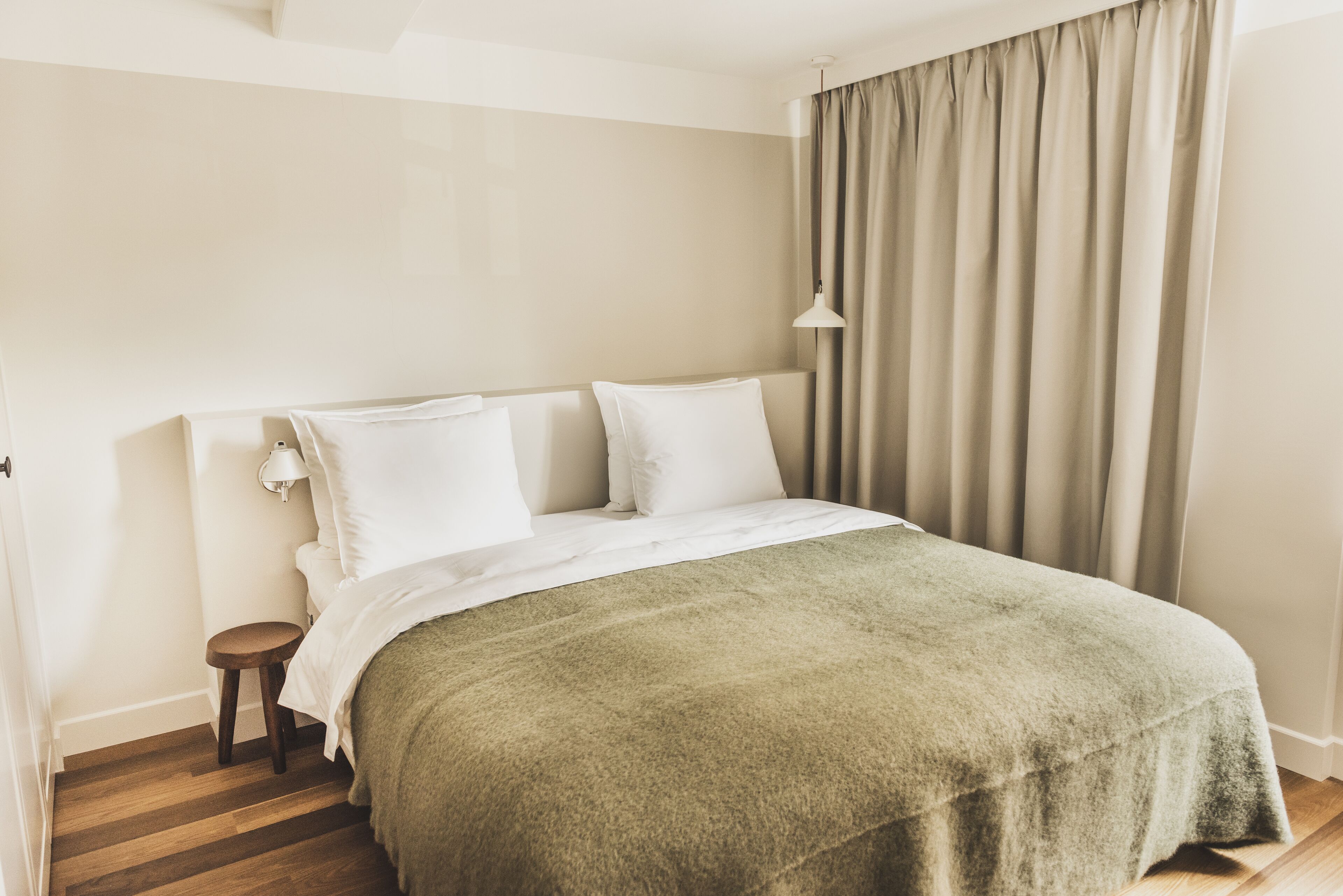 A neatly made bed with a green blanket, a small wooden stool, and a bedside lamp.