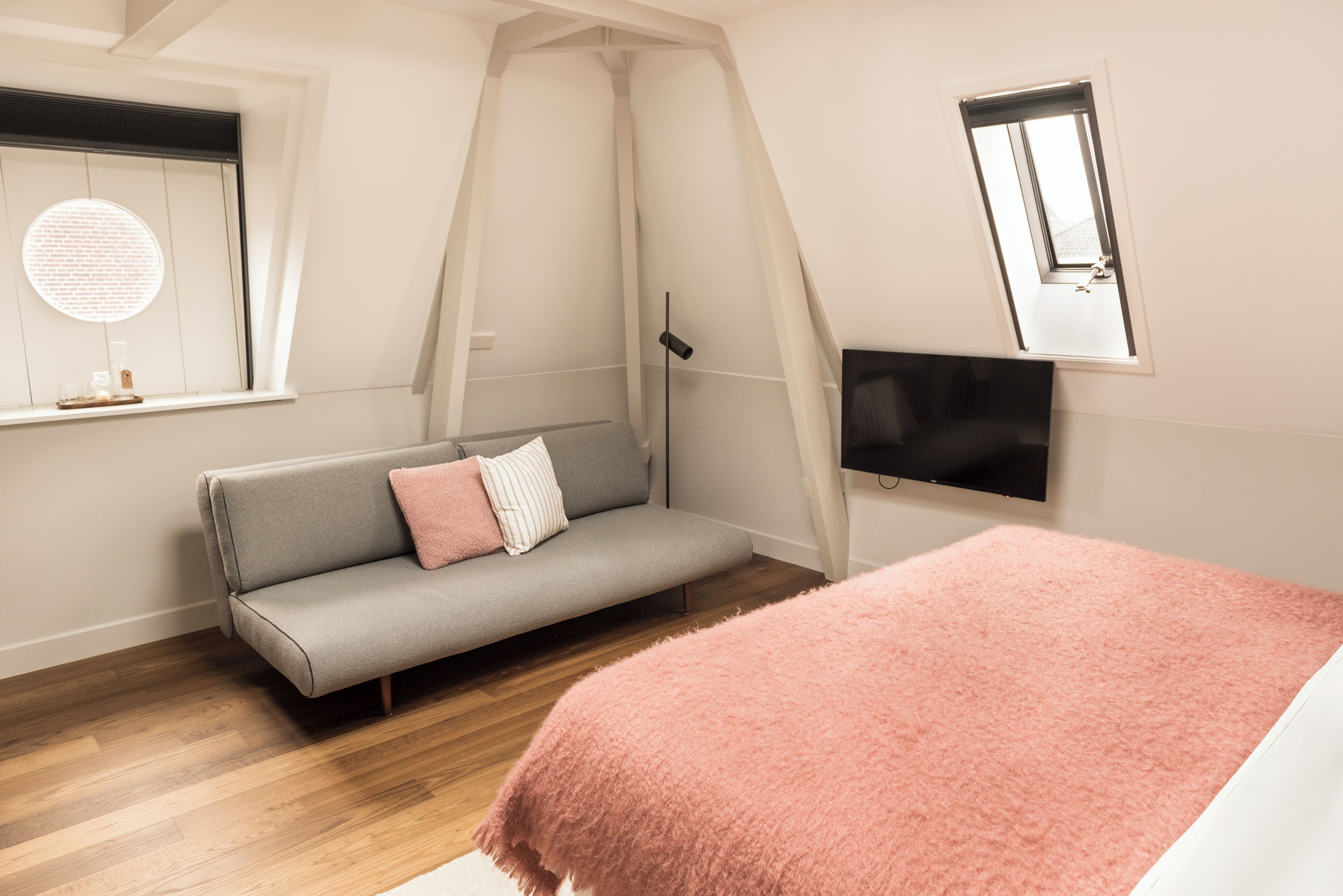 A bedroom with a grey sofa, a pink blanket, a TV, and a window with a curtain.