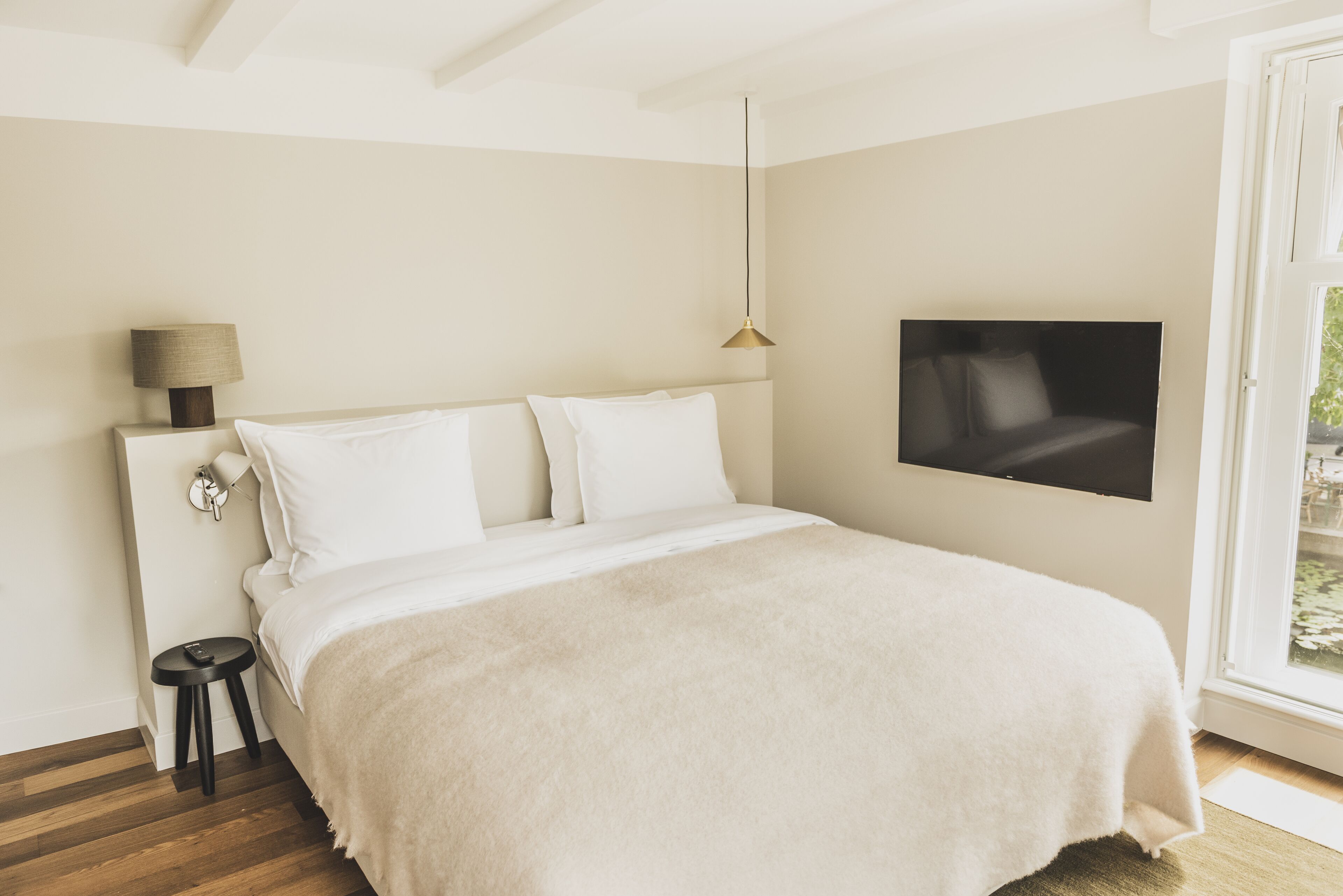 A bedroom with a bed, a TV mounted on the wall, a lamp on a bedside table, and a window with a view.