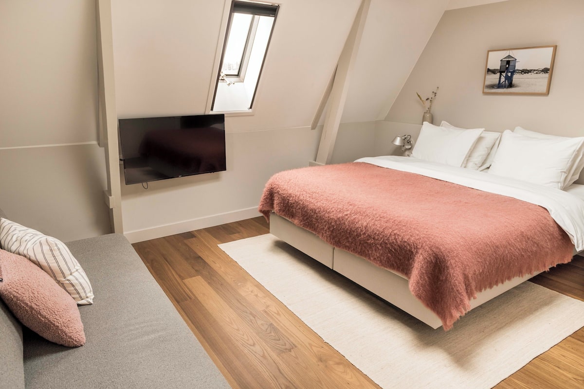 A bedroom with a bed, a TV mounted on the wall, a window, and a painting on the wall.