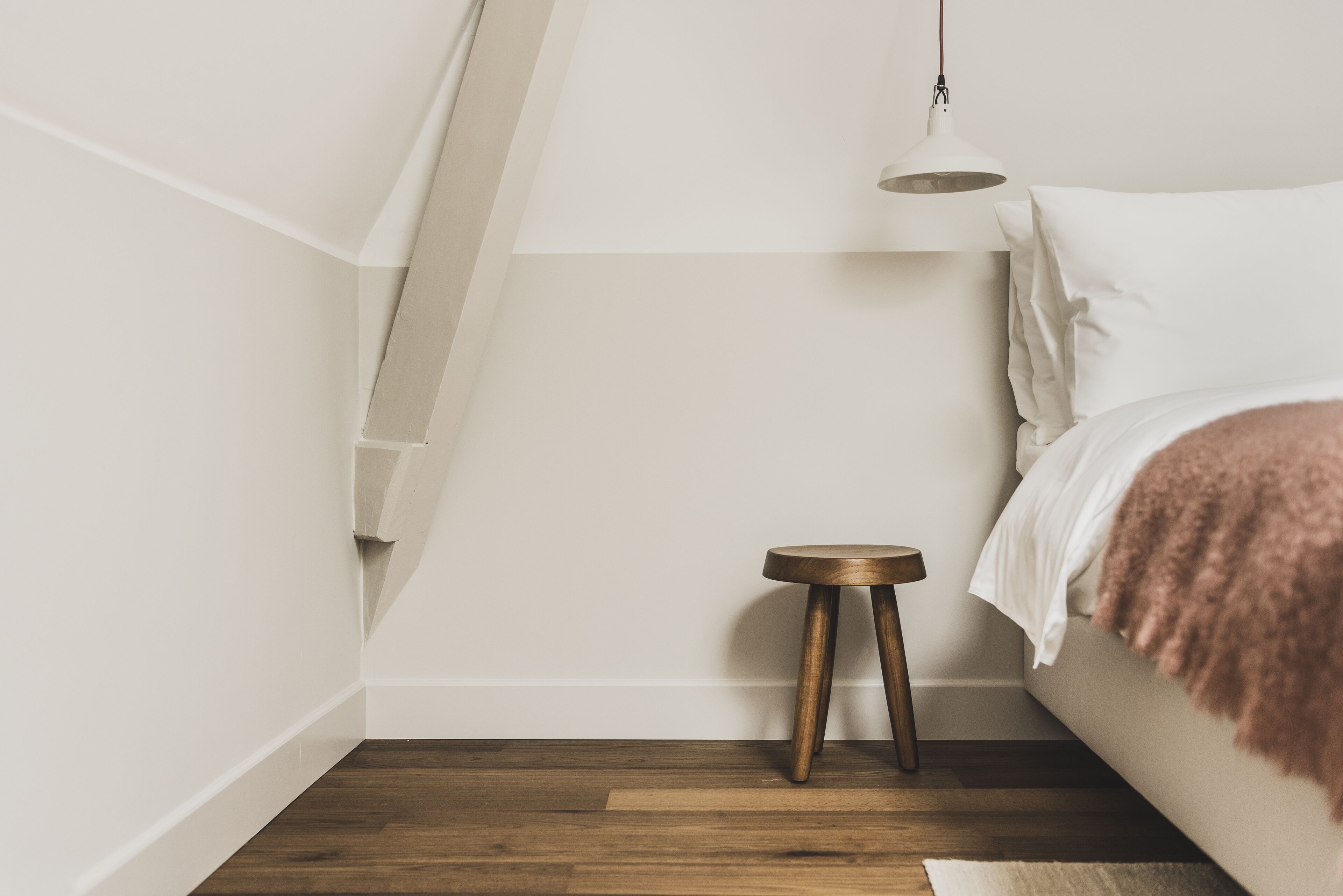 A bedroom with a bed, a small wooden stool, a white lamp, and a sloped ceiling.
