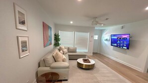 A modern living room with a beige sofa, a glass coffee table, and a flat-screen TV mounted on the wall. - Culdesac Heights Cottage with Enclosed Parking (Houston)