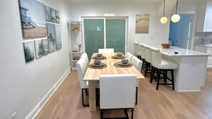 A modern dining area with a wooden table and white chairs, a kitchen with white cabinets, and a view of the outdoors through a glass door. - Culdesac Heights Cottage with Enclosed Parking (Houston)